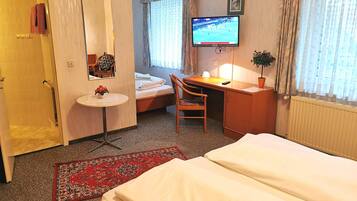 A hotel room with a bed, a desk, a chair, a small table with a vase, a TV, and a window with curtains.