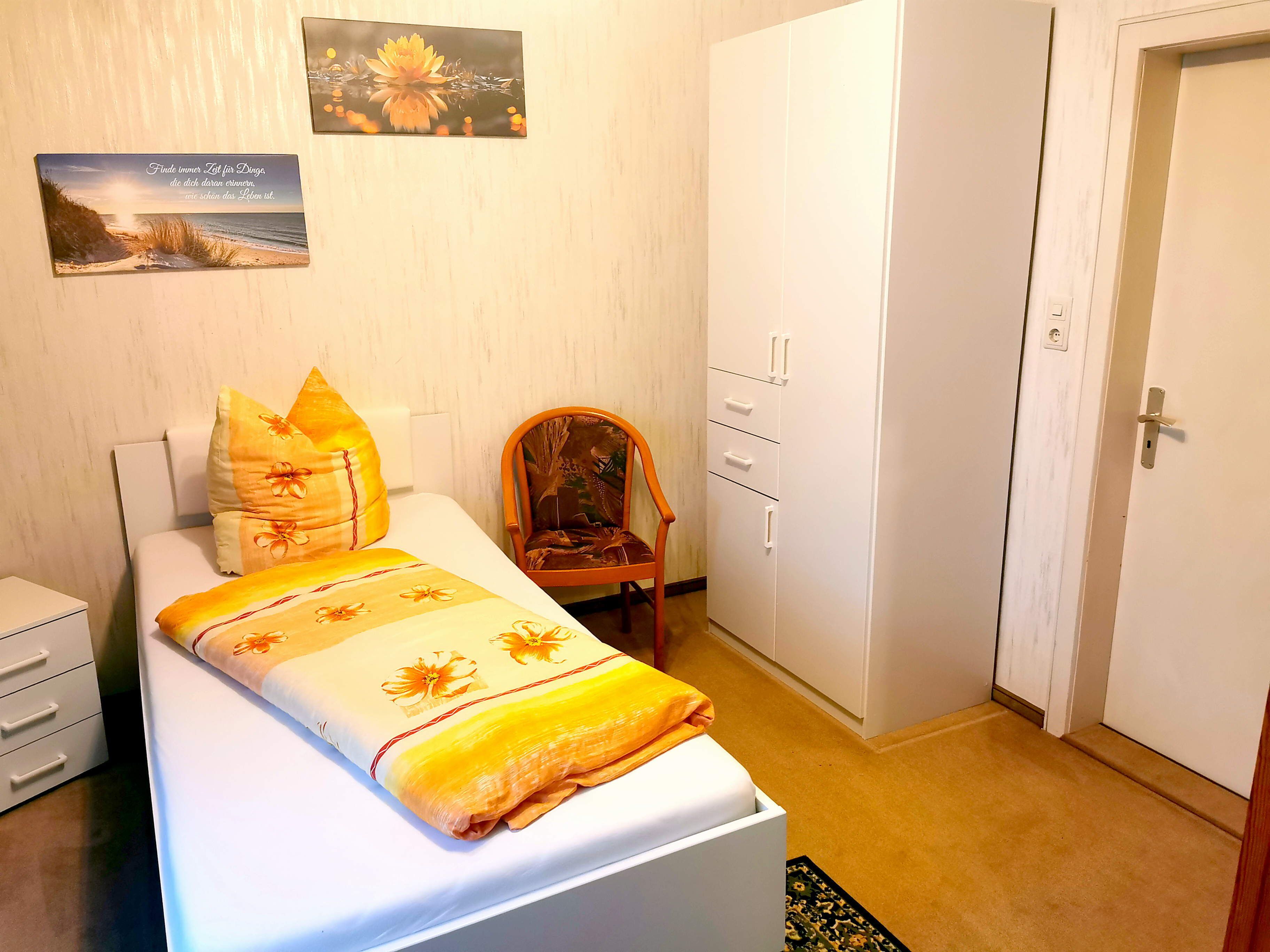 Un dormitorio ordenado con una cama, una silla, un armario y dos cuadros colgados en la pared.