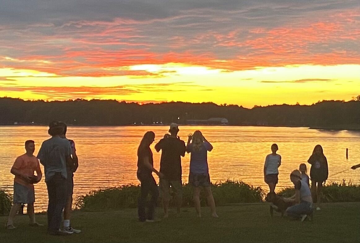 A group of people standing by a lake during sunset.
