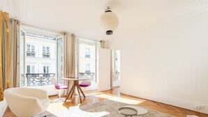 A spacious living room with a large window, a round wooden table, and a white armchair.
