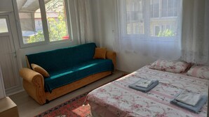 A bedroom with a sofa, a bed with floral bedding, a window with curtains, and a view of greenery outside. - Tan Apart Aile Ev Pansiyonu (Yesilova)