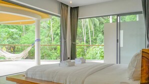 Ein Schlafzimmer mit einem großen Bett, einem hölzernen Kopfteil und Blick auf üppiges Grün durch große Fenster.