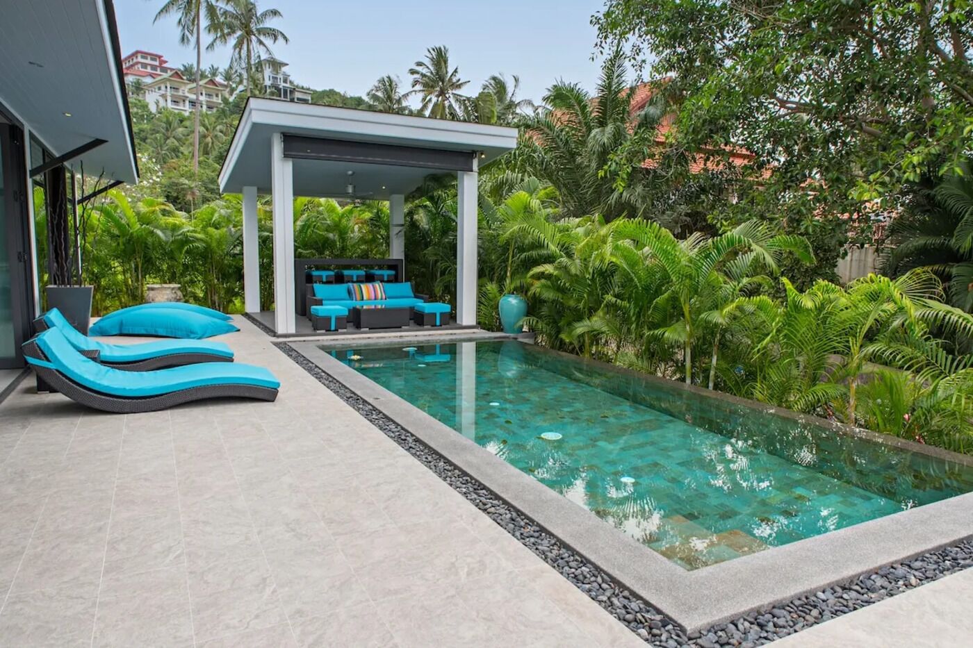 Ein Swimmingpool mit Liegestühlen und einer überdachten Sitzgruppe, umgeben von üppiger Vegetation.