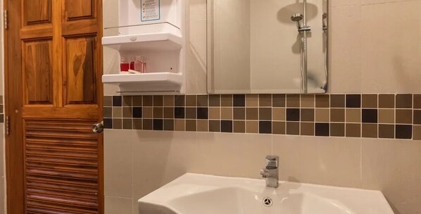 Une salle de bain avec une porte en bois, un lavabo blanc, une douche et un mur recouvert de carreaux derrière le lavabo.