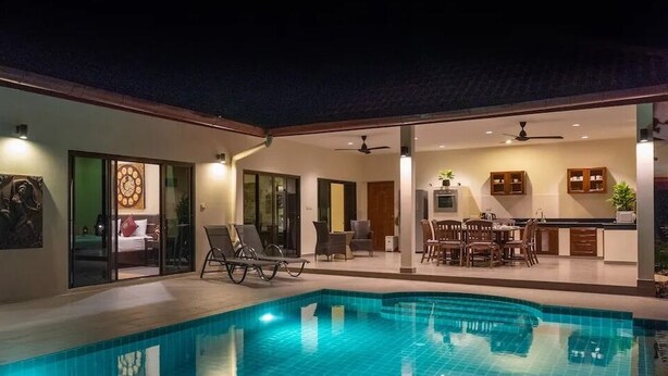 Une piscine avec des chaises longues, un coin repas et une cuisine visible à travers des portes vitrées.