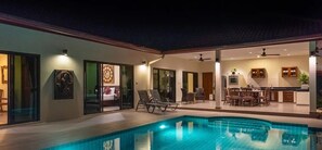 Une piscine avec des chaises longues, un coin repas et une cuisine visible à travers des portes vitrées.