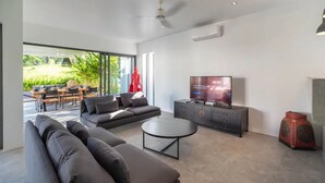 A modern living room with a large sofa, a coffee table, a TV, and a cabinet. There is a large window providing a view of the outdoors. - Elegant 4-bedroom villa with private pool, waterfall showers, and garden (Tambon Mae Nam)