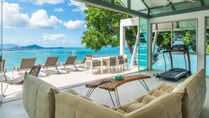 Une chambre moderne dotée d’une grande baie vitrée donnant sur une plage et l’océan. On y trouve un tapis de course, une table basse en bois et un canapé beige.