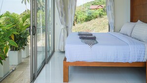 Une chambre dotée d’un grand lit, d’une porte-fenêtre coulissante donnant sur un balcon, et offrant une vue sur une colline verdoyante avec un bâtiment.