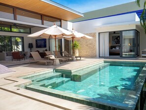 Un espace piscine moderne avec des chaises longues et des parasols, une piscine carrelée en pierre et une pièce fermée par des baies vitrées offrant une vue panoramique.