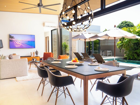 A modern dining area with a pool view, a ceiling fan, and a TV displaying a beach scene.
