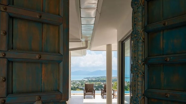 A view from inside a building looking out to a balcony with a chair and a table, overlooking a landscape with mountains and a body of water.
