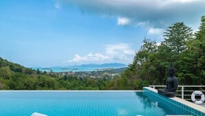 A infinity pool with a Buddha statue, overlooking a coastal town and hills.