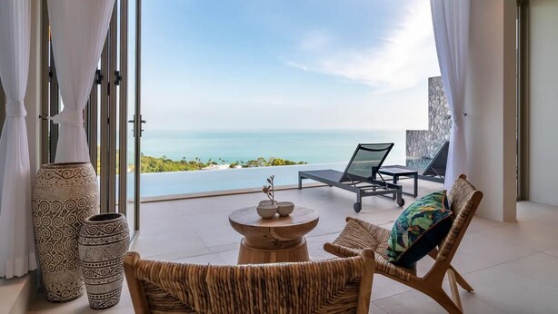 A modern room with a large glass door leading to a balcony overlooking the sea. There are wicker chairs, a small round table, and decorative vases. - Luxurious 3 king-suite villa in Plai Laem Soi 8 with private pool, sea views, and WiFi (Koh Samui)