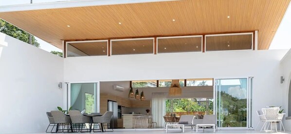 A modern house with a dining area, kitchen, and large sliding glass doors leading to a patio. - Luxurious 4-bedroom villa near Chaweng Beach with private pool, mountain views, and WiFi (Tambon Bo Put)