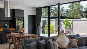 A modern living room with a grey sofa, a dining area with wooden chairs, and large windows with a view of palm trees. - Modern 2-bedroom oasis with private pool, garden, and dedicated workspace, ideal for families (Tambon Mae Nam)
