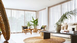 A modern living room with a sofa, coffee table, and chairs. There are large windows with sheer curtains, and a potted plant on the coffee table.