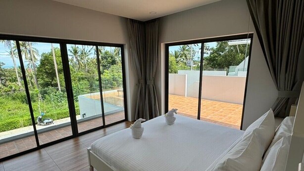 A modern bedroom with a large bed, a white bedside table, and a view of a garden and building through large windows. - Stylish 2-Bedroom VIlla in Plai Laem, Private Pool with Jacuzzi, Close to Attractions (Koh Samui)