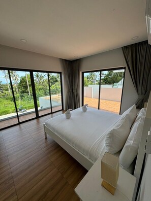 A modern bedroom with a large bed, a white bedside table, and a view of a garden and building through large windows.
