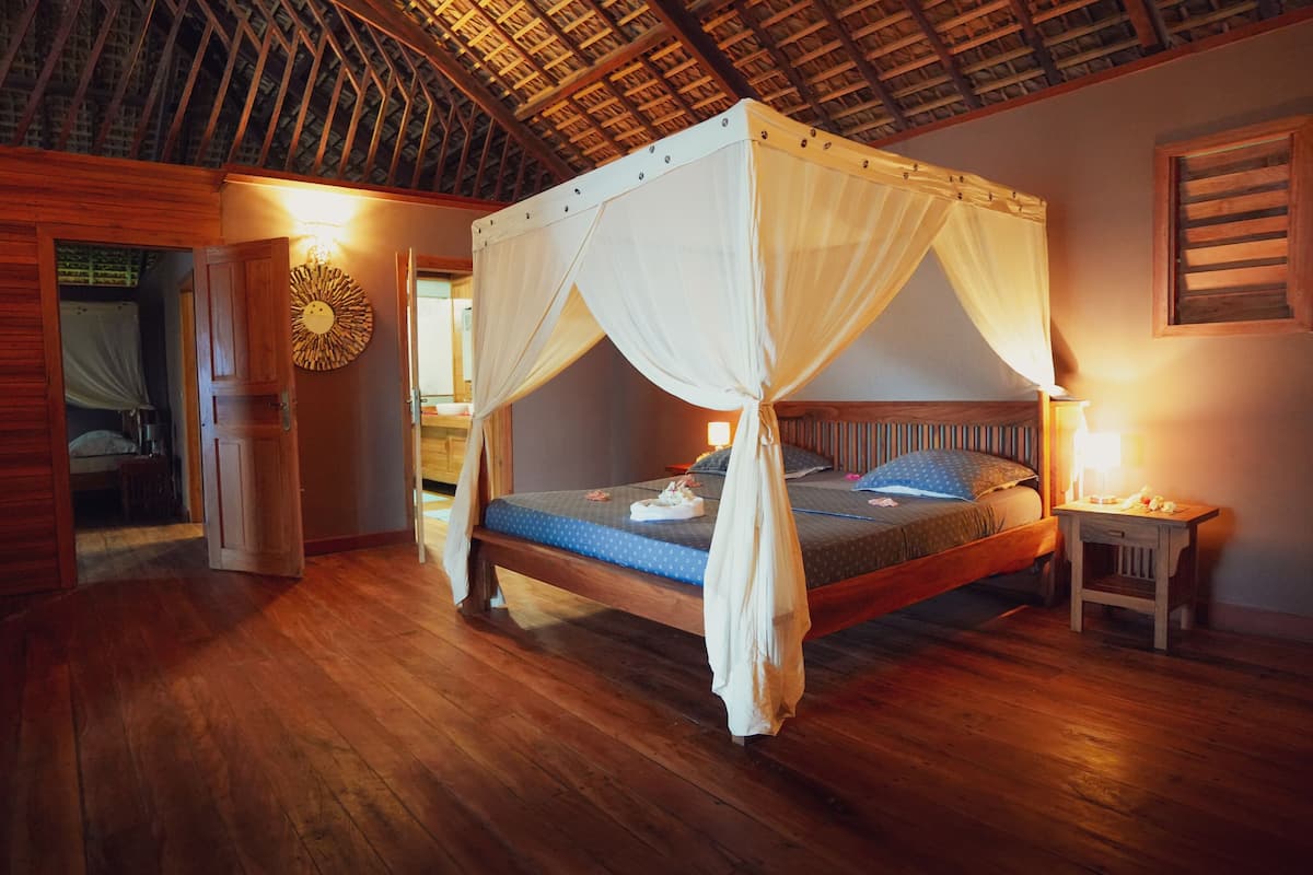 Uma cama de dossel com estrutura de madeira, uma mesa de cabeceira com abajur e um relógio fixado na parede.