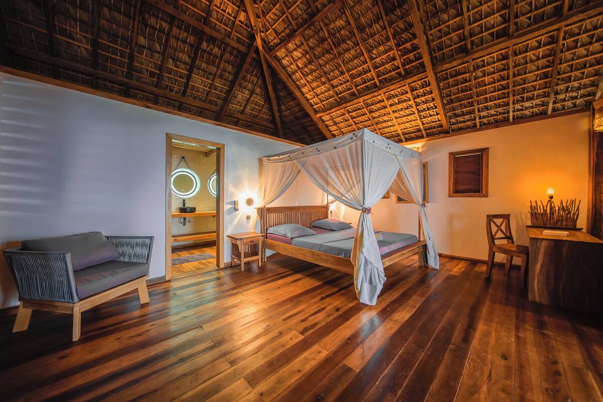 Um quarto com piso de madeira, uma cama com dossel, um sofá, uma mesa pequena e uma cadeira.