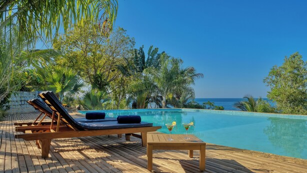 Una terraza de madera con sillones de descanso y una mesa, con vistas a una piscina azul cristalina y al océano.
