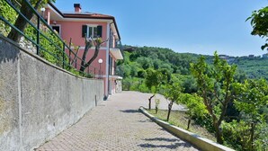 Ein gepflasterter Weg führt zu einem rosafarbenen Gebäude mit einem Balkon, umgeben von Grünflächen und Hügeln.