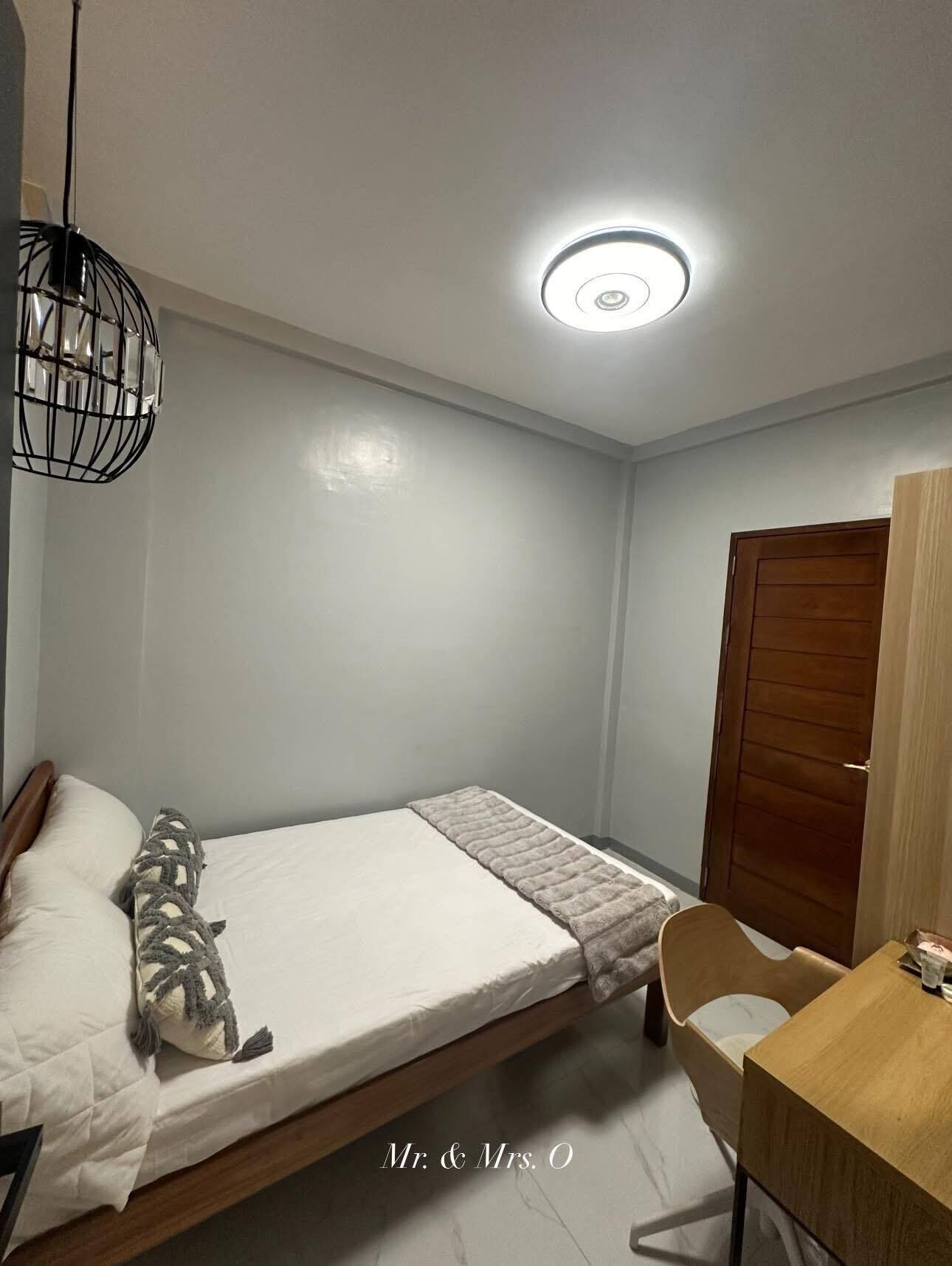 Ein Schlafzimmer mit einem Bett, einem Holztisch, einem Stuhl, einer Hängelampe und einer Tür.