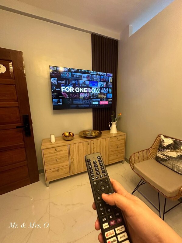 Un salon avec un téléviseur à écran plat fixé au mur, qui diffuse une publicité pour une offre promotionnelle, un meuble en bois orné d’objets décoratifs, un canapé et une télécommande à portée de main.