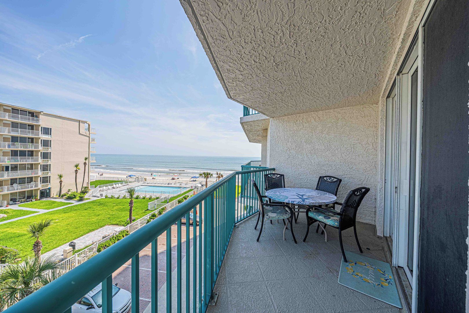 A balcony with a table and chairs, overlooking a beach and a pool.