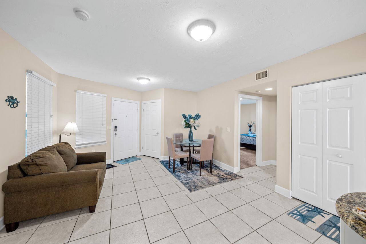 A living room with a brown sofa, a dining table with chairs, and a kitchen area with a white door.