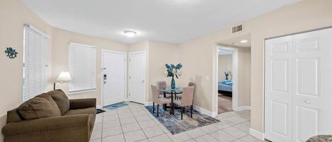 A living room with a brown sofa, a dining table with chairs, and a kitchen area with a white door.