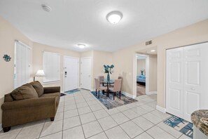 A living room with a brown sofa, a dining table with chairs, and a kitchen area with a white door.