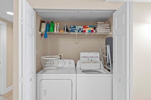A laundry room with two washing machines, a drying rack, and shelves above containing cleaning supplies and folded towels.
