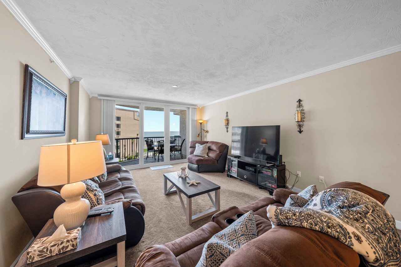 A living room with a sofa, a coffee table, a TV, and a balcony with outdoor furniture.