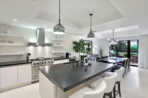 Une cuisine moderne dotée d’un îlot central, d’appareils électroménagers en acier inoxydable, de luminaires suspendus et offrant une vue sur l’extérieur.