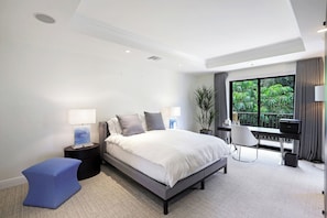 A modern bedroom with a large bed, bedside lamps, a small table, a chair, and a view of greenery through the window.