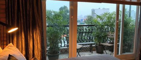 Un dormitorio con una cama grande, vistas de la ciudad desde un balcón y una planta en maceta en el balcón.