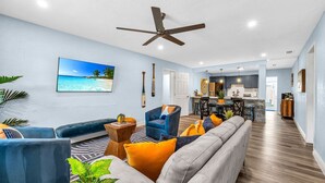 A spacious living room with a large sofa, a blue armchair, and a dining area with a kitchen in the background.