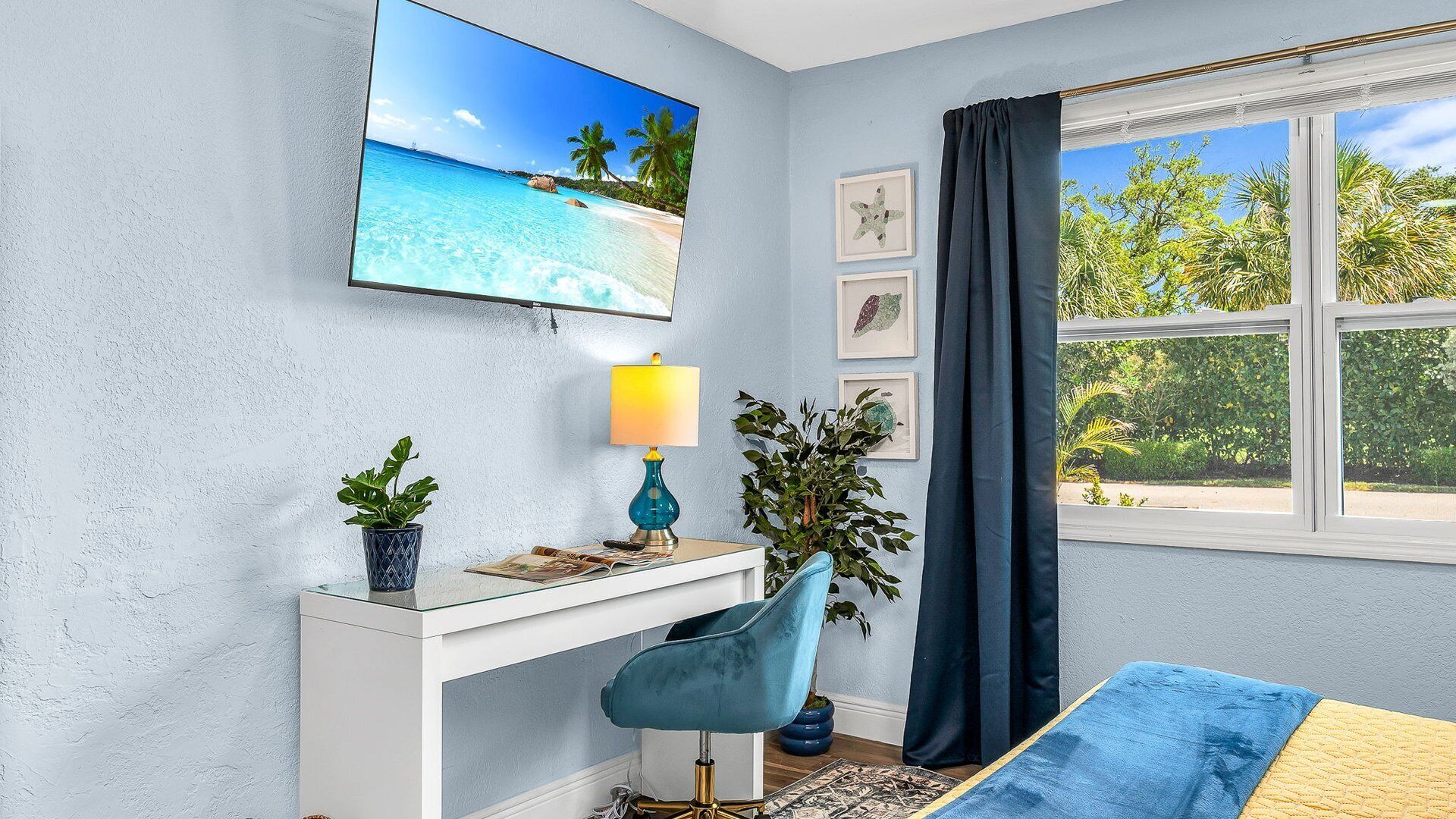 A room with a desk, a chair, a lamp, a bed, a window with curtains, and a wall with framed pictures.