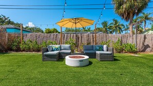 A backyard with a grassy lawn, two wicker sofas with cushions, a round fire pit, and a yellow umbrella.