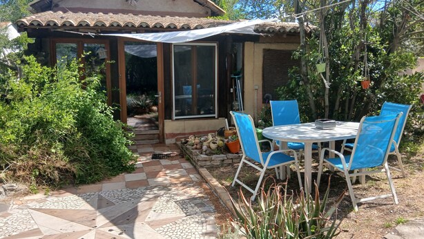 Une terrasse avec une table et des chaises, un chemin dallé et une maison avec un toit en tuiles et de grandes fenêtres.