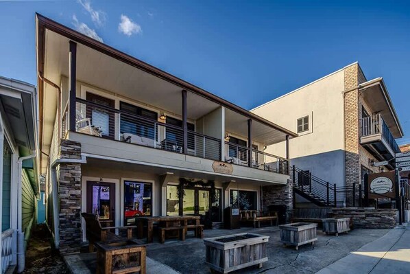 Um edifício de dois andares com telhado plano e varanda. Possui áreas externas com mesas e bancos.