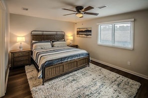 Quarto com cama, duas mesas de cabeceira, ventilador de teto, janela com persianas e um tapete.