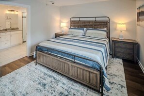 A bedroom with a large bed, wooden headboard, two bedside tables with lamps, a nightstand, and a bathroom visible in the background.