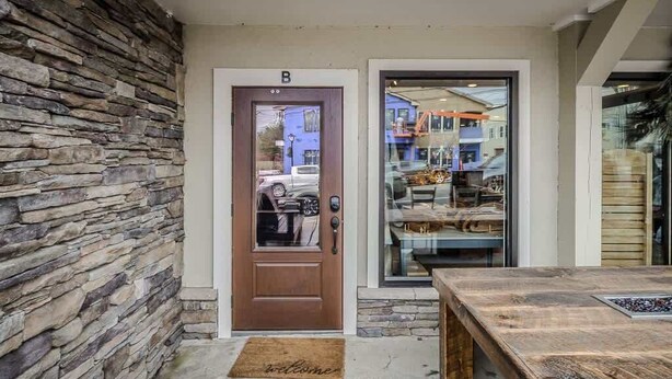 A stone wall, a wooden door with a 'B' above it, and a wooden table with a welcome mat in front.