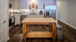 A modern kitchen with white cabinets, a wooden table, stainless steel appliances, and a small trash bin.