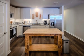 A modern kitchen with white cabinets, a wooden table, stainless steel appliances, and a small trash bin. - BR Walkabout B - Steps to Main Street restaurants, (Blowing Rock)