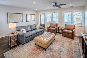 A living room with a grey sofa, a brown leather ottoman, and two leather chairs. There is a patterned rug on the floor, a small table with a lamp, and a ceiling fan. - BR Walkabout C - Steps to Downtown Blowing Rock - (Blowing Rock)