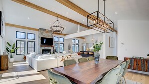 Un vaste salon doté d’une grande table à manger en bois, de sièges confortables, d’une cheminée en pierre et, au fond, d’une cuisine moderne.